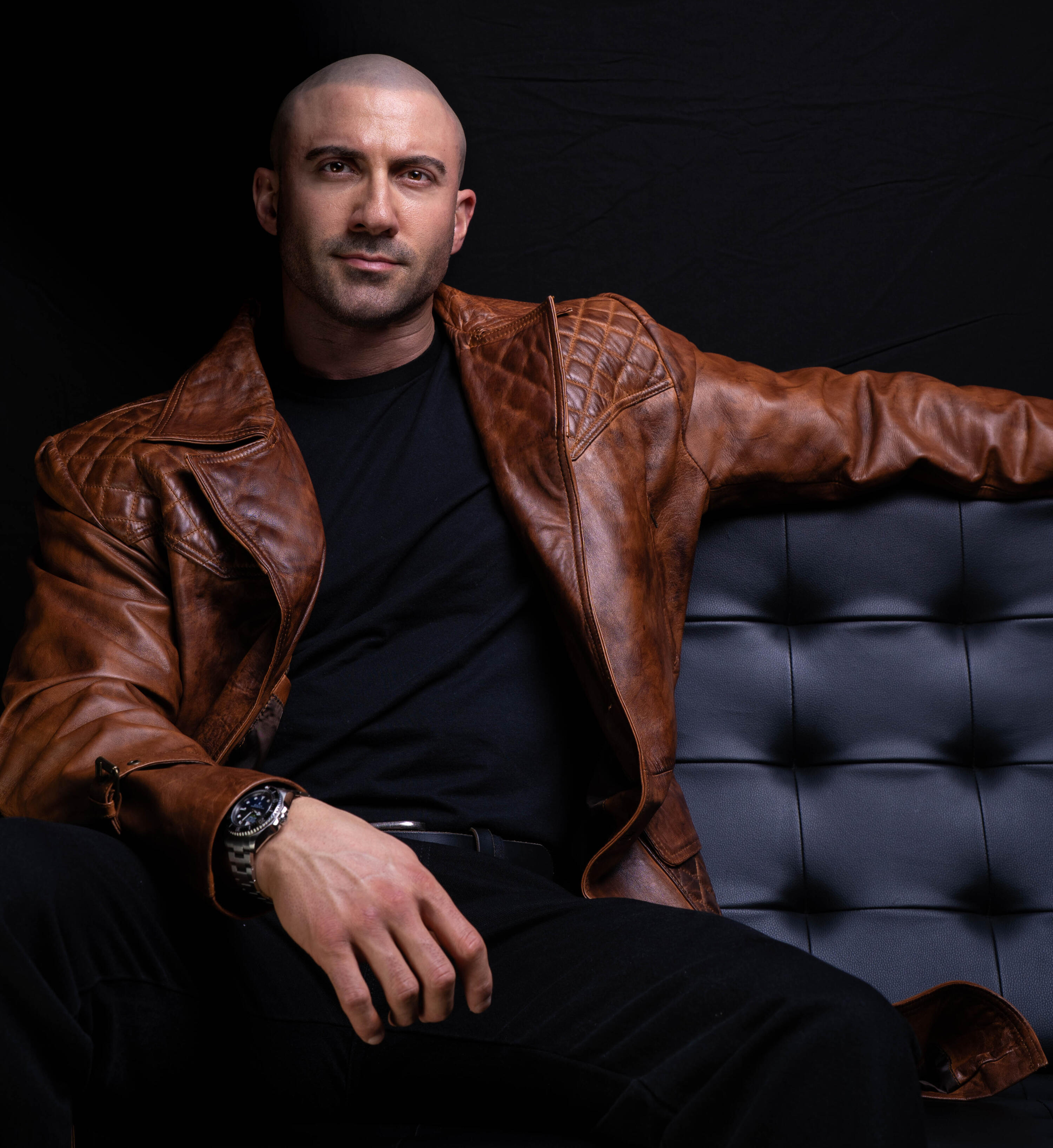 Tom Rooney, Author, Founder, Headline Performer Author Thomas Rooney (Tom Rooney), sitting in a black Barcelona chair wearing long brown leather blazer, promoting Reflections of a Male Revue Headliner: How to Win at Life and Love